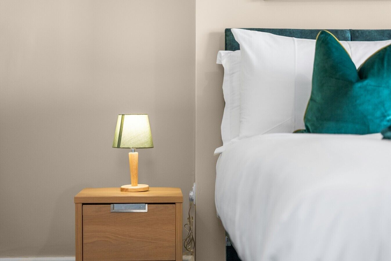 Un lit bien fait, avec une literie blanche et un coussin turquoise, une table de chevet avec une lampe et une petite table en bois.