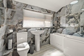 Een badkamer met toilet, wastafel en badkuip, alles in het wit, tegen marmerlook wanden.