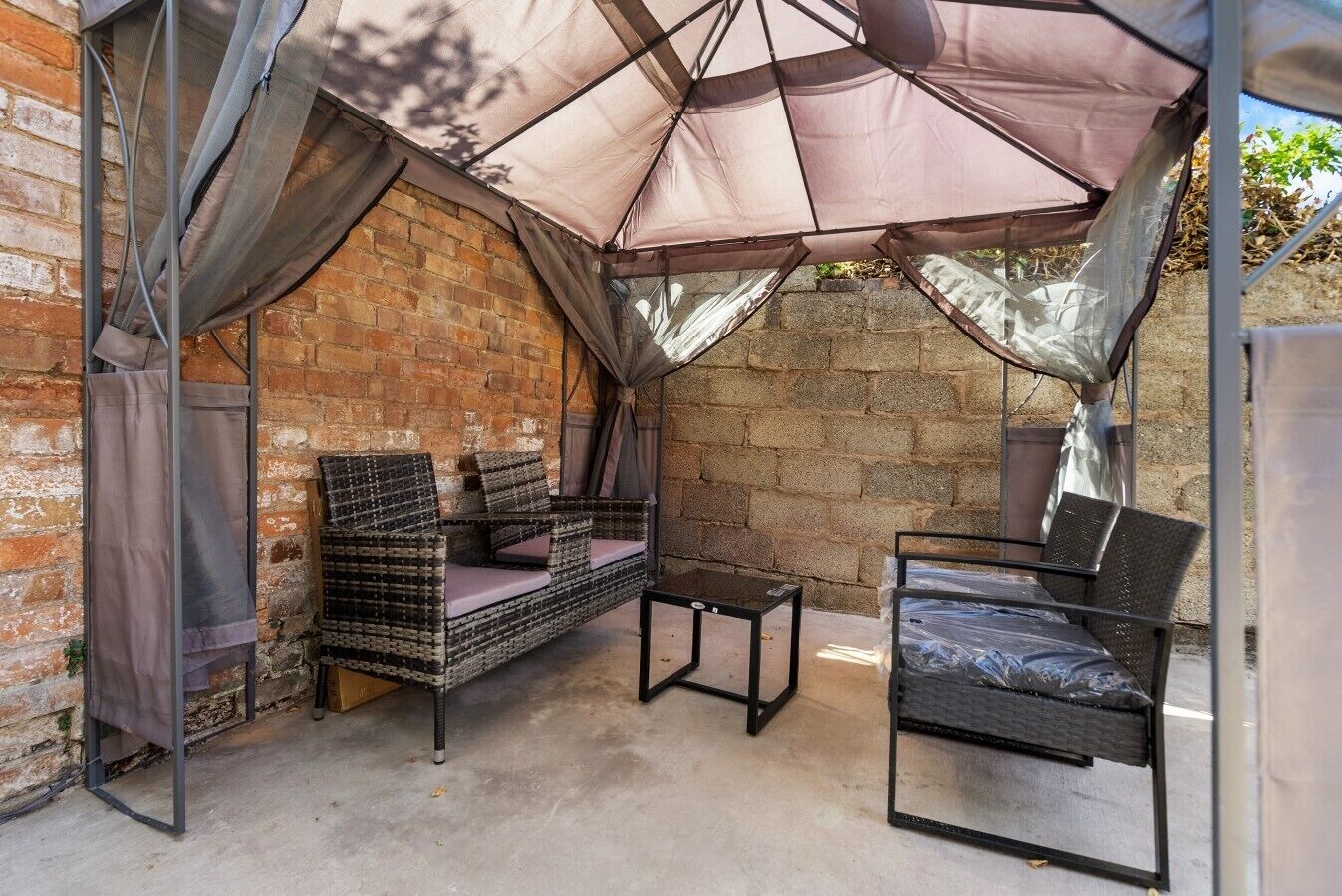 A covered outdoor seating area with a wicker sofa and a metal table, set against a brick wall.