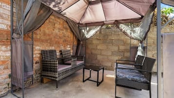 A covered outdoor seating area with a wicker sofa and a metal table, set against a brick wall.