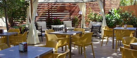 An outdoor patio with yellow chairs and tables, a covered seating area, and a wooden fence in the background.