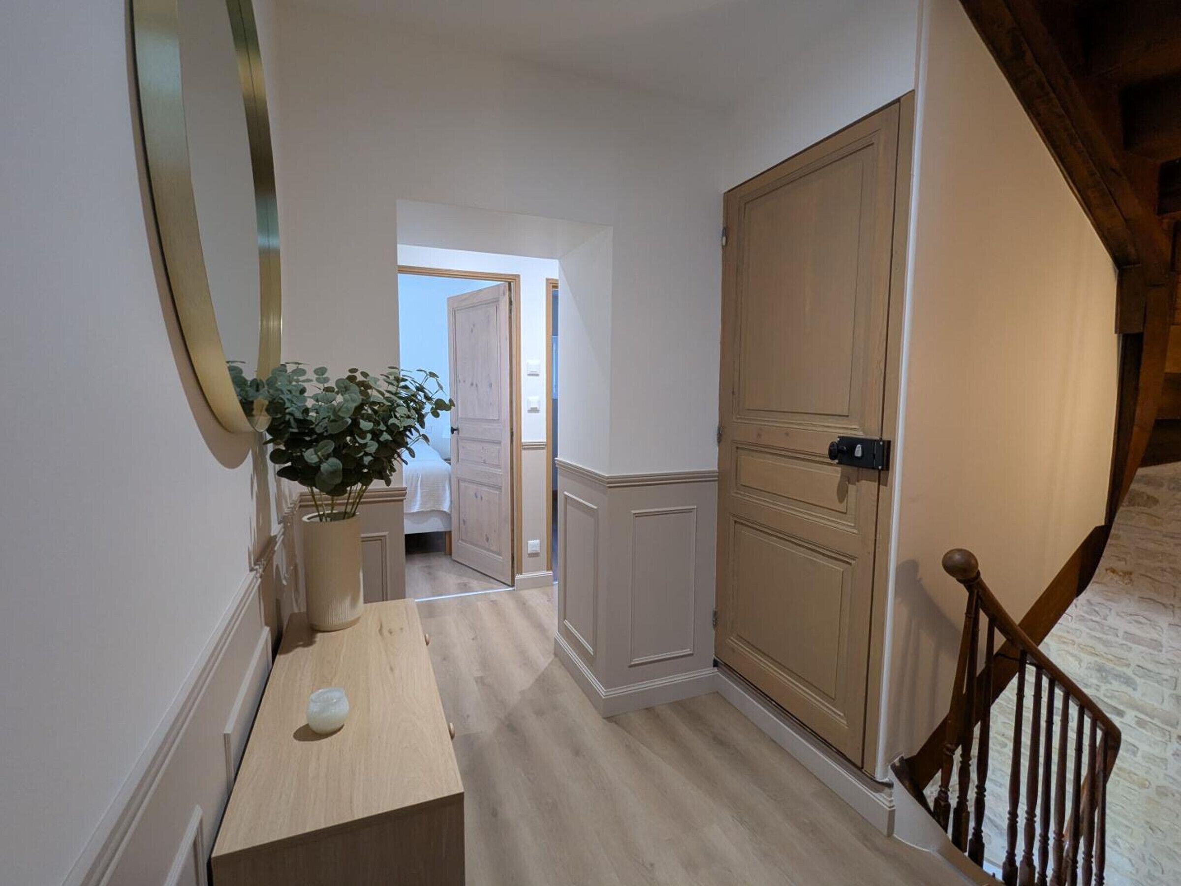 A hallway with a wooden floor, a potted plant, a mirror, and a door leading to another room.