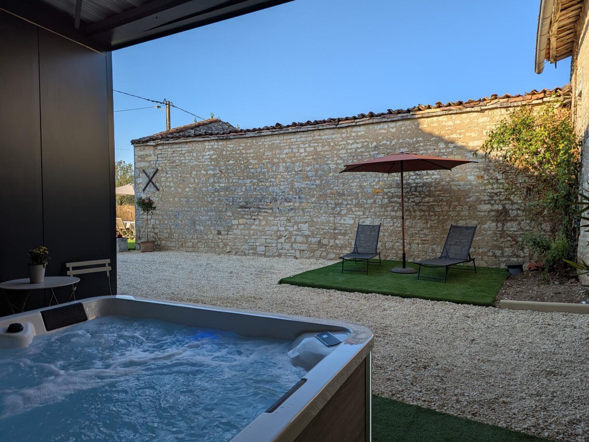 A hot tub in a covered outdoor area with a stone wall, two chairs, and an umbrella.