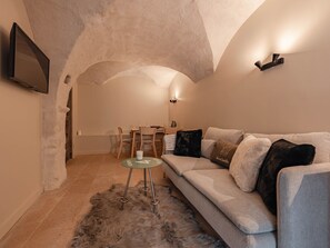 Ein gemütliches Wohnzimmer mit einer hohen, gewölbten Decke, einem grauen Sofa mit vielen Kissen, einem kleinen Glastisch und einem flauschigen Teppich sowie einem Essbereich mit Stühlen und Tisch.