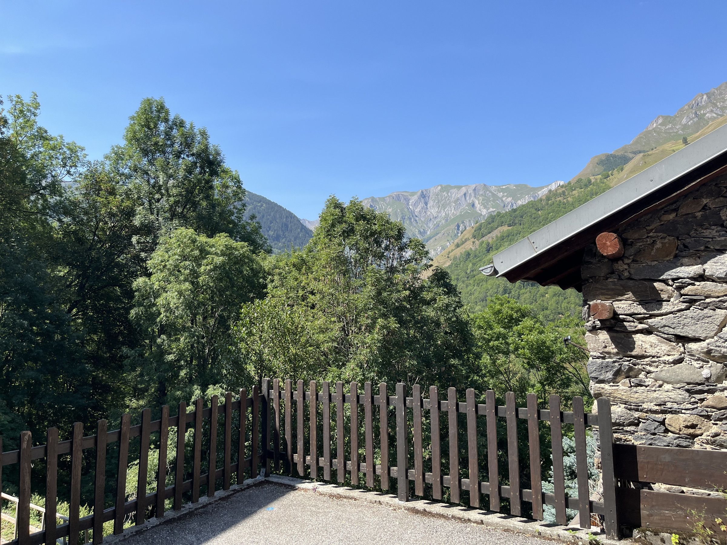 Un edificio de piedra con una cerca de madera al frente, rodeado de árboles y montañas al fondo.