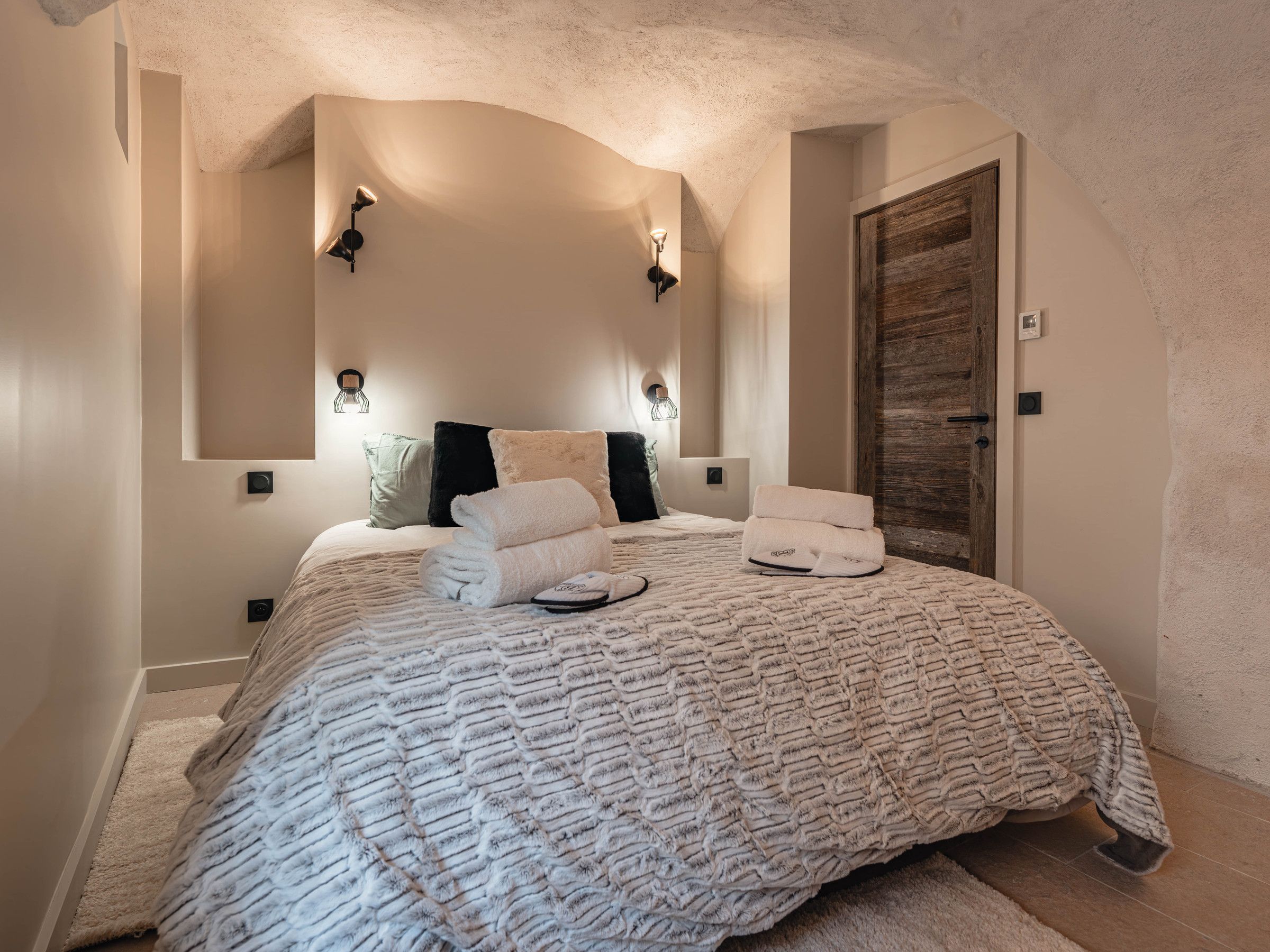 A bedroom with a large bed, a wooden door, and wall-mounted lighting.