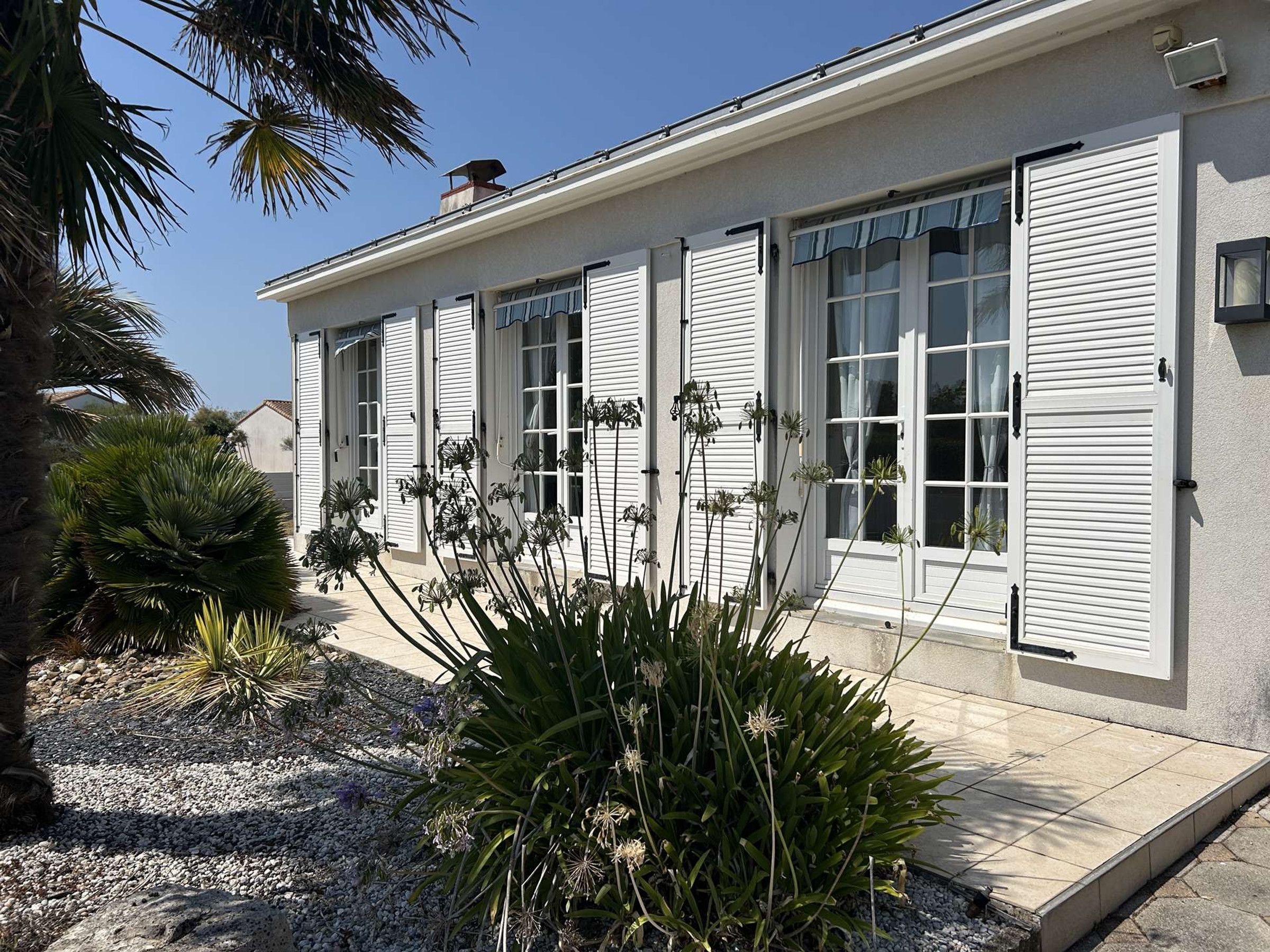 A building with white shutters, a tiled patio, and a garden with plants.