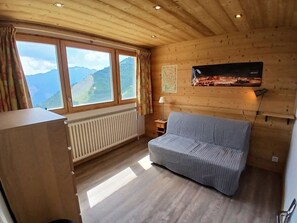 Ein Zimmer mit Holzboden, einem Sofa, einem Heizkörper, einem fenster mit Vorhängen und einem an der Wand befestigten Fernseher.