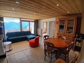 A room with a wooden ceiling, a sofa, a red chair, a dining table with chairs, and a view of mountains through large windows. - Apartment in Vieil Alpe - 6 guests (Huez)