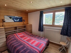 Ein Schlafzimmer mit einem Bett, einem an der Wand montierten Fernseher, einem Fenster mit Vorhängen, einem Heizkörper und einem Sessel.