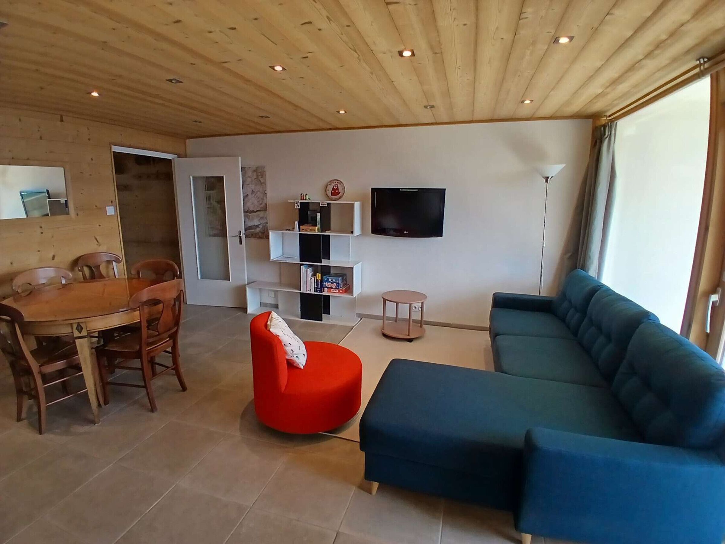 A living room with a wooden ceiling, a dining table with chairs, a red ottoman, a blue sofa, a TV on the wall, and a bookshelf.
