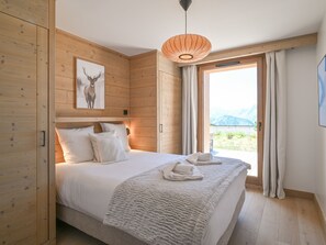 A bedroom with a bed, a wooden headboard, a wall-mounted picture of a deer, and a view of mountains through a window. - Elegant apartment, panoramic view, mountain - 7 guests (Huez)