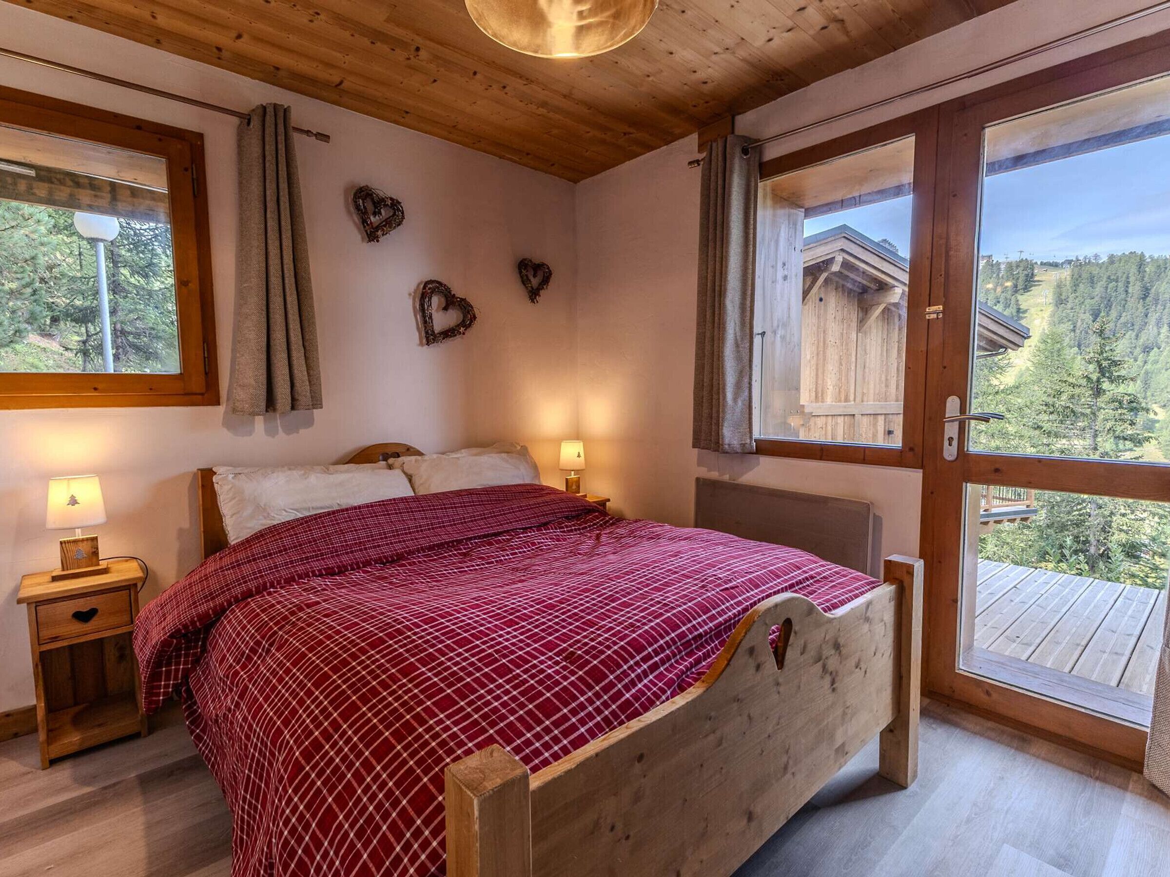 A bedroom with a wooden bed, two bedside tables, a window with curtains, and a view of a wooden cabin.