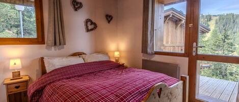 A bedroom with a wooden bed, two bedside tables, a window with curtains, and a view of a wooden cabin.