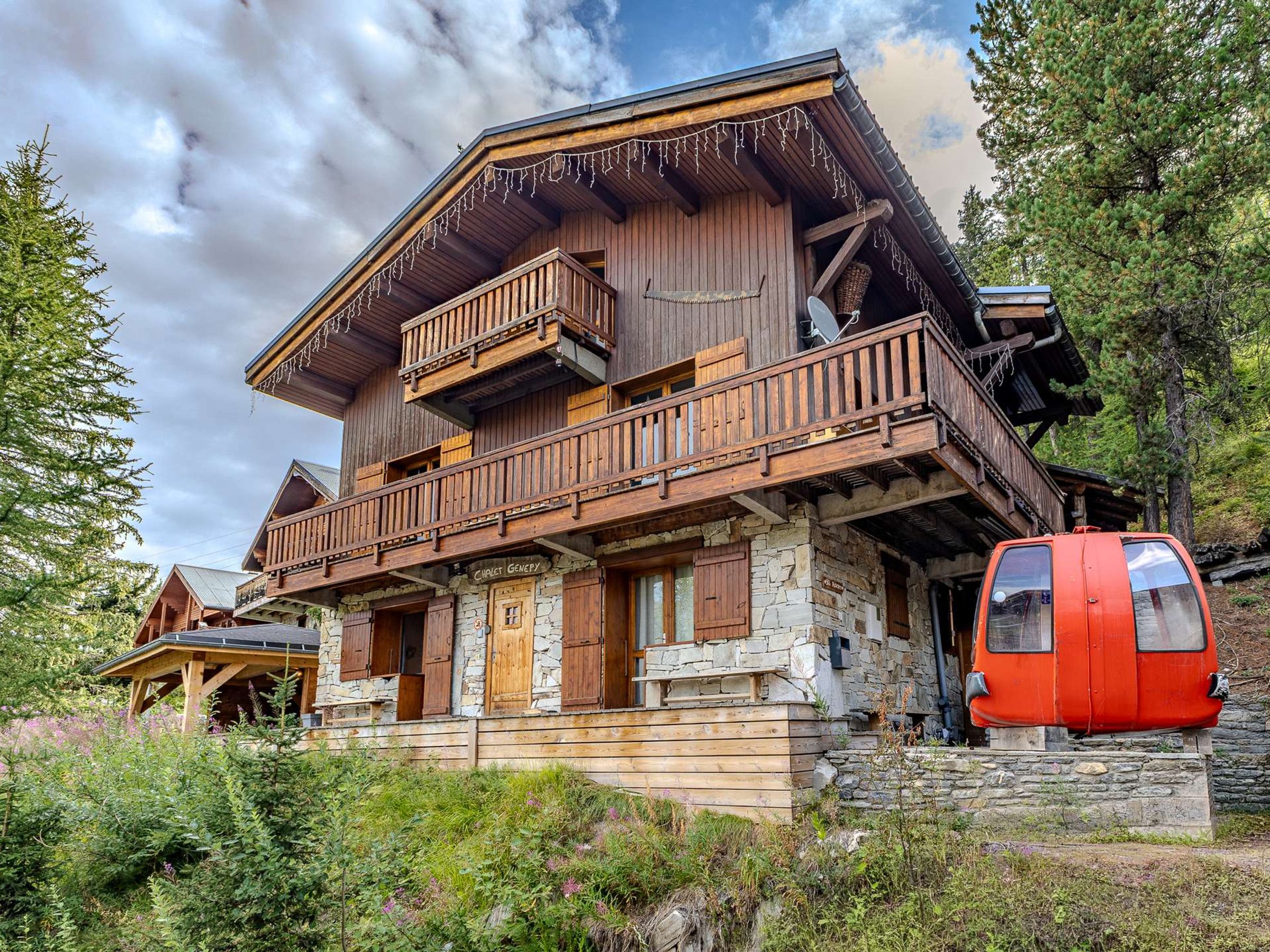 A wooden chalet with a stone foundation, wooden balconies, and a red cable car in front.