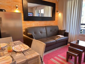 Ein möbliertes Wohnzimmer mit einer Couch, einem Esstisch mit Tellern und Besteck, einem Fernseher und einem Kühlschrank.