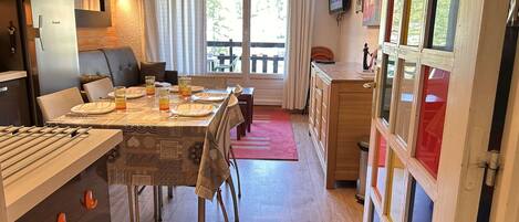 A dining area with a table set for four, a kitchen with a refrigerator, and a window with curtains.