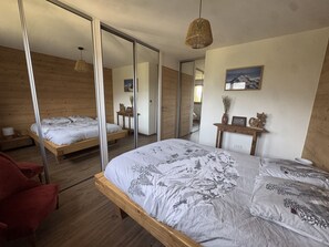 Ein Schlafzimmer mit einem großen Bett, Holzboden, einem roten Sessel, einem Nachttisch mit einer Vase und einem Spiegel an der Wand.