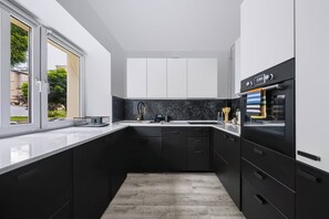 Eine moderne Küche mit schwarzen Schränken, einer weißen Arbeitsplatte, einem eingebauten Backofen und einem Fenster mit Blick ins Freie.