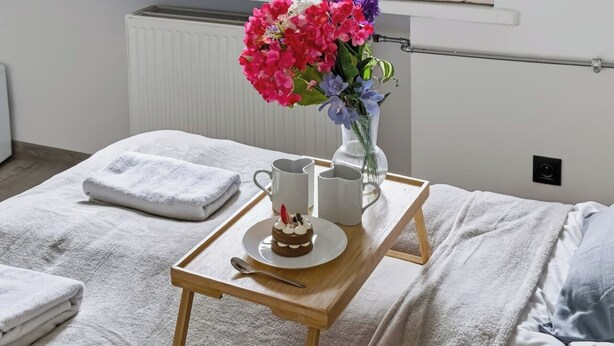 En snyggt bäddad säng med ett brickbord som står på den, med en kaka, två muggar och en vas med blommor.