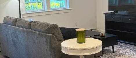 Un salon avec un canapé gris, une petite table ronde surmontée d’un vase vert, un meuble TV noir et une fenêtre avec des stores.