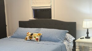 A bedroom with a bed, a bedside table with a lamp, a window with a blind, and a wall-mounted heater.