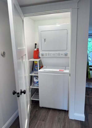 A compact laundry room with a washing machine and dryer, a shelf with cleaning supplies, and a door leading to another room. - Beautiful studio apartment with AC in South Kingstown, Rhode Island. (South Kingstown)