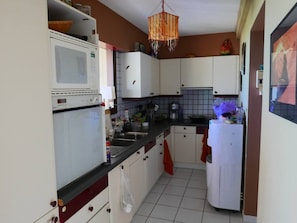 A compact kitchen with white cabinets, a microwave, a refrigerator, and a hanging light fixture. - Charming 100-square-meter apartment + 40-square-meter terrace with a stunning view of the sea and lagoon (Saint-Paul)