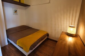 Una cama individual con colchón marrón y manta amarilla, una mesita de noche de madera con lámpara, y una pared blanca con un cuadro pequeño.