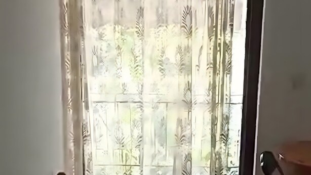 Ein Einzelbett mit Holz Kopfteil, ein kleiner Tisch und Blick auf einen Balkon durch transparente Vorhänge.