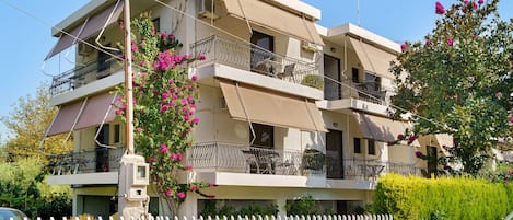 A multi-story residential building with balconies, a white fence, and a car parked in front.