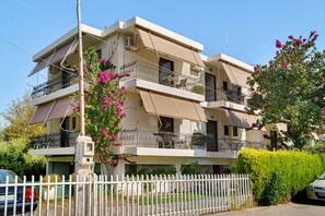 A multi-story residential building with balconies, a white fence, and a car parked in front. - Rousetis Two-Bedroom Apt 205 - 200m from the Sea (Aigialeia)