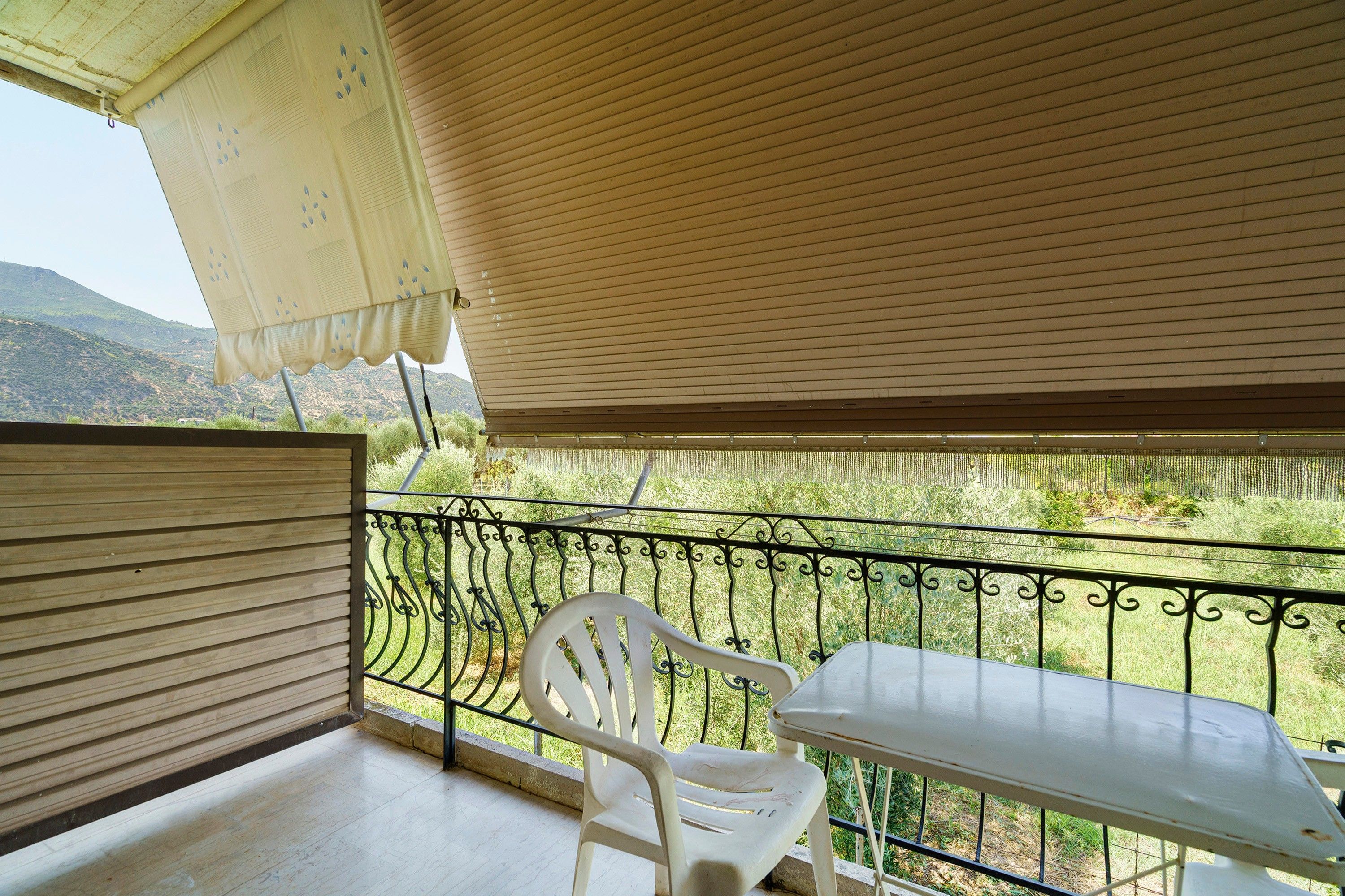 Un balcón con una silla y mesa de plástico blanco, barandilla metálica y vista a un paisaje montañoso.