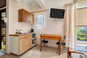 A compact kitchen with wooden cabinets, a sink, and a small table with chairs. A flat-screen TV is mounted on the wall, and a window with curtains is visible. - Rousetis Studio 104 - 200m from the Sea in Aígio (Aigialeia)