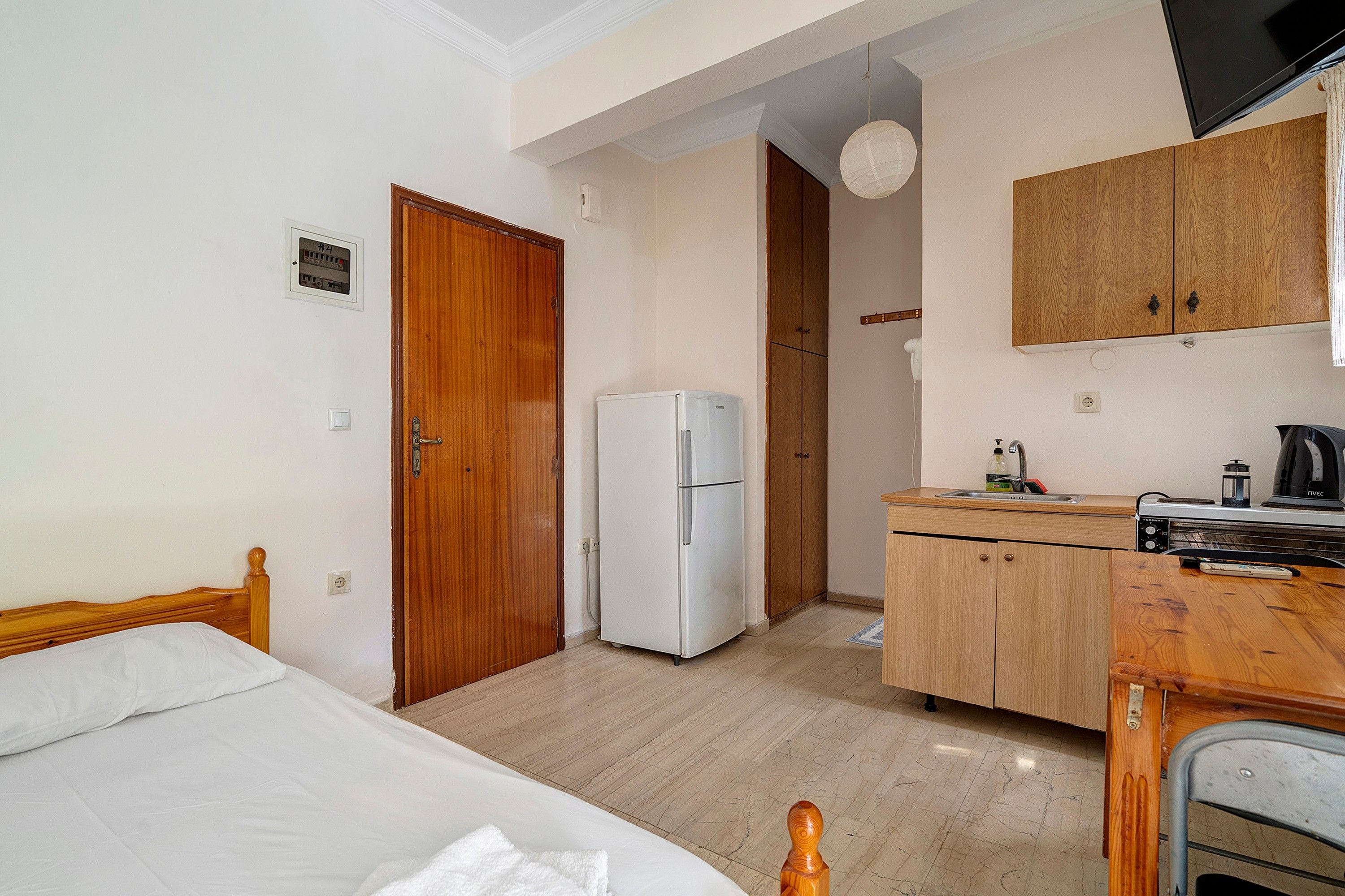 A compact hotel room with a bed, a kitchenette, a refrigerator, and a wooden wardrobe.