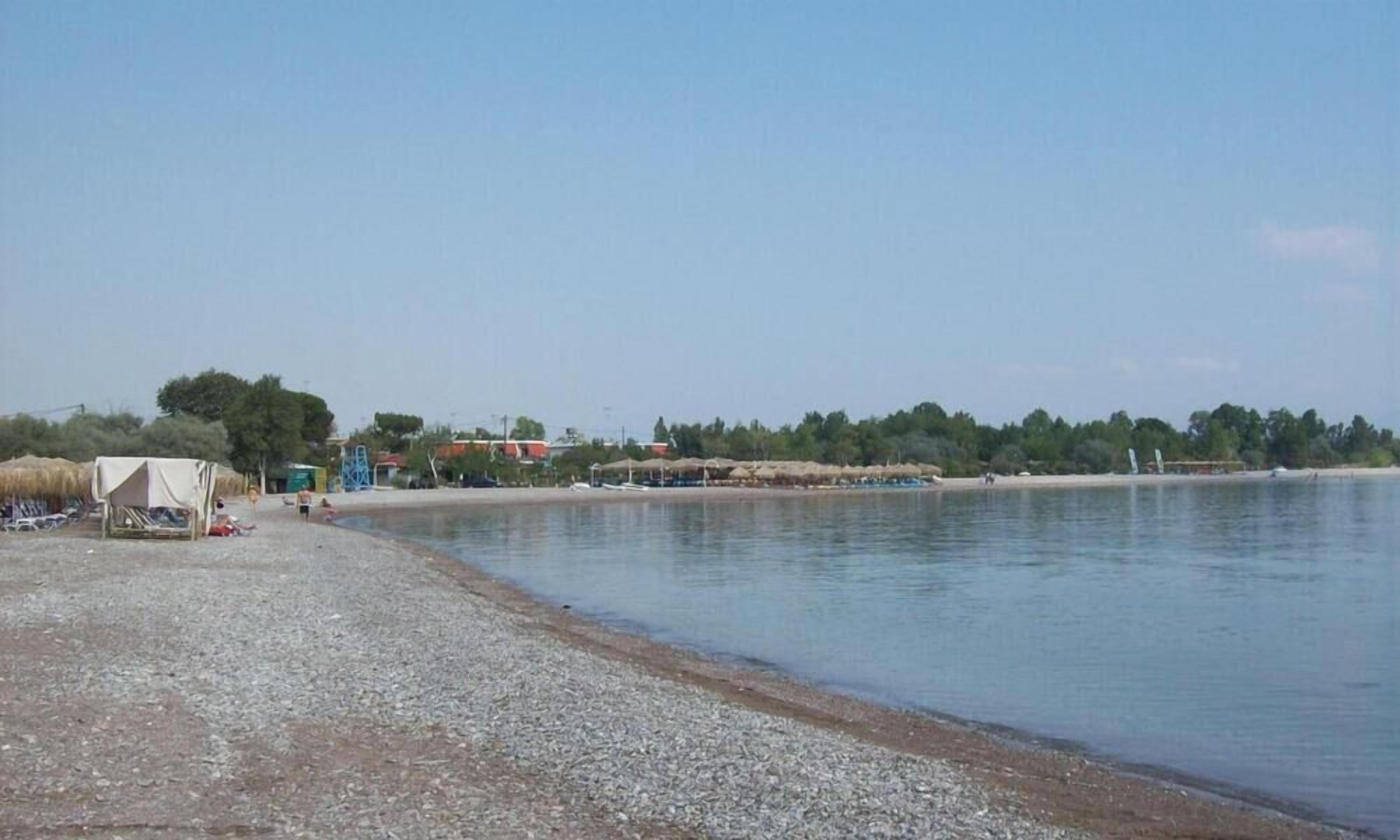 En strand med ett tält och en liten byggnad, stilla vatten och träd i bakgrunden.