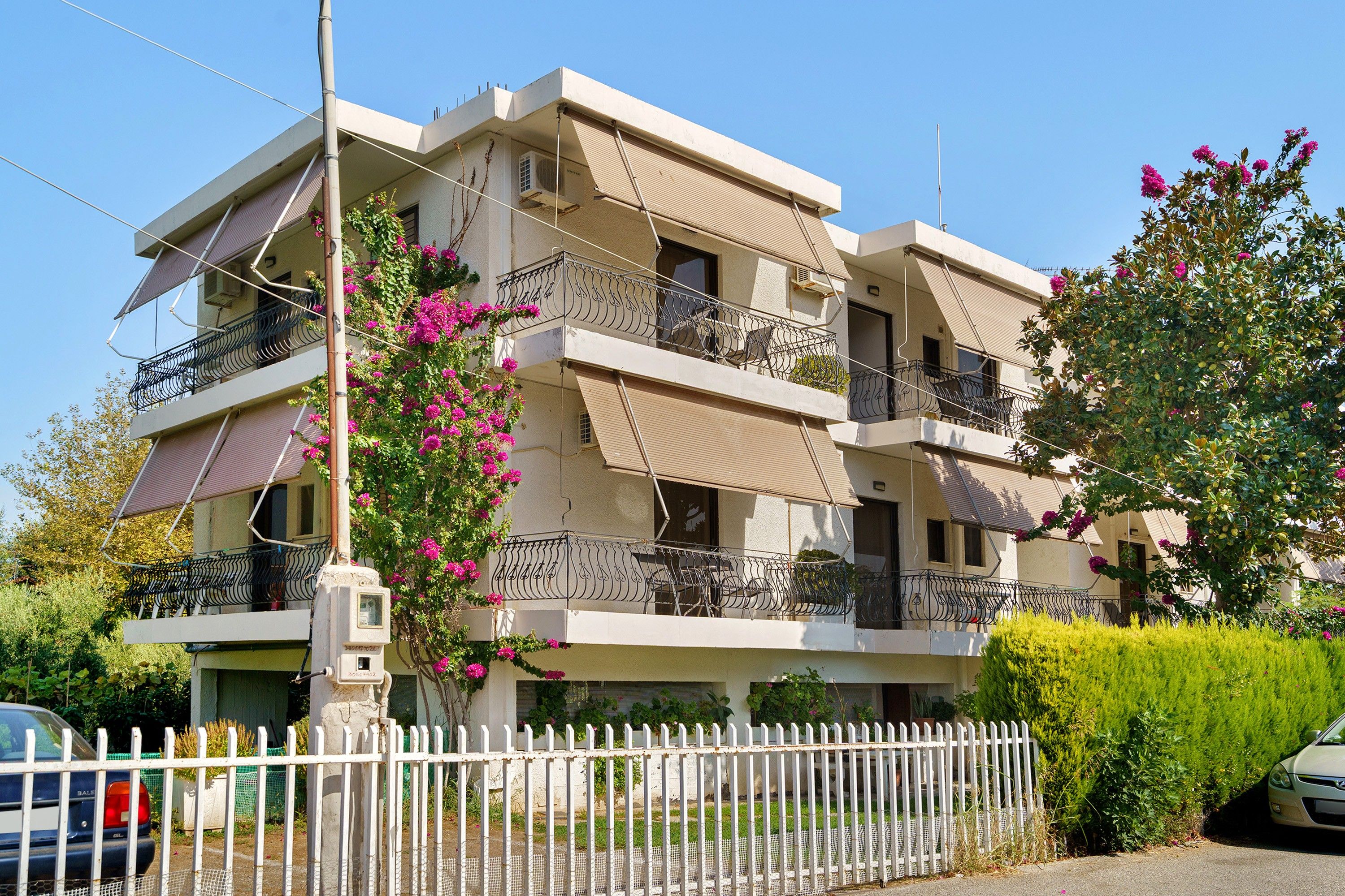 A multi-story residential building with balconies, a white fence, and a car parked in front.