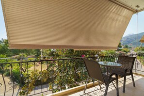 A balcony with a table and chairs, a retractable awning, and a view of a garden and distant hills. - Rousetis Two-Bedroom Apt 203 - 200m from the Sea (Aigialeia)