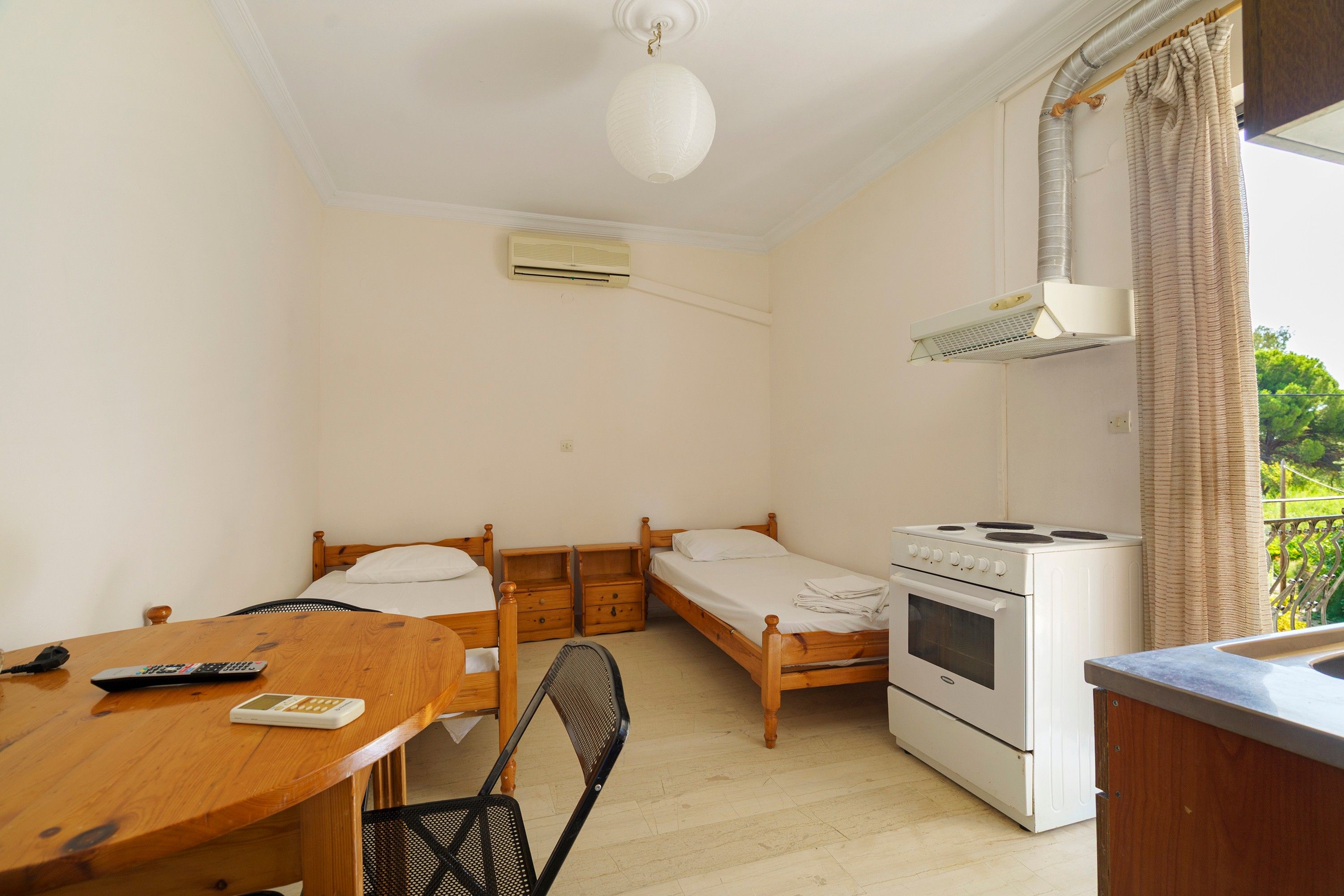 Una habitación pequeña y limpia con dos camas individuales, una mesa de madera con una silla, una estufa blanca y un ventilador de techo.