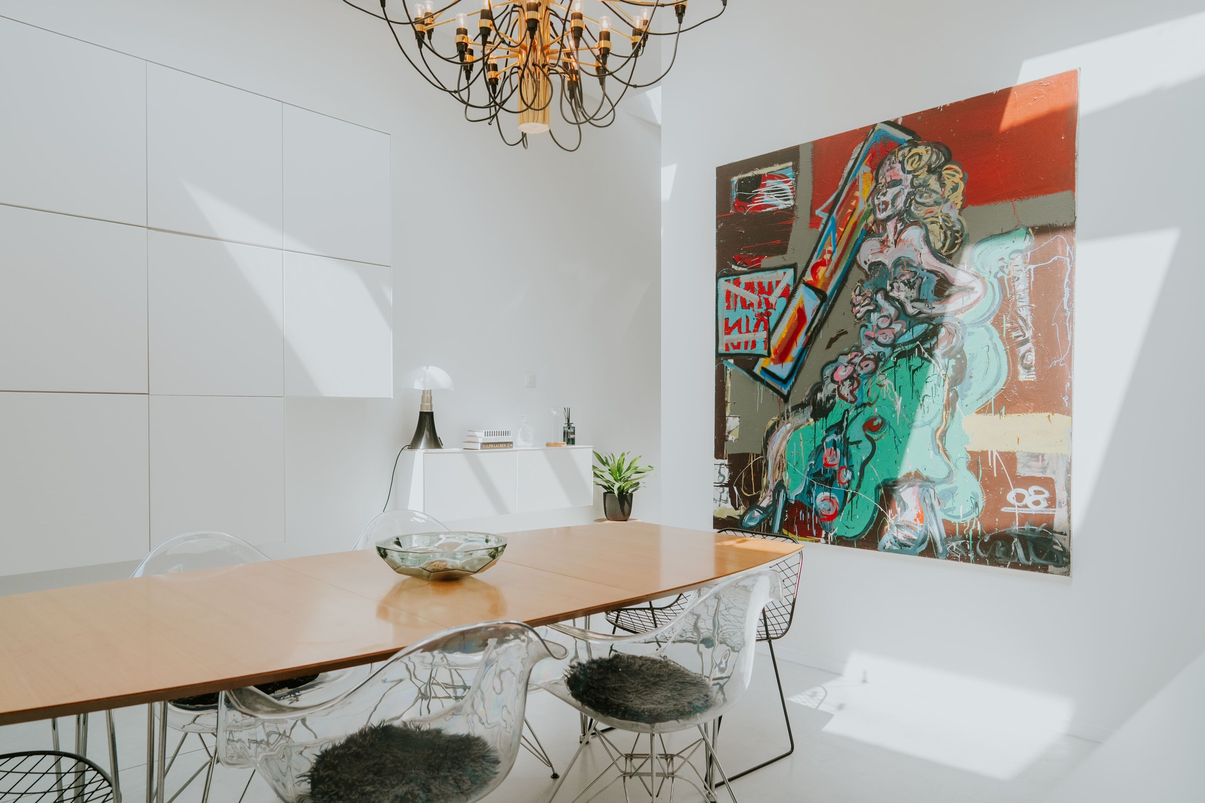 A modern dining area with a wooden table, clear glass chairs, a chandelier, and a large abstract painting on the wall.