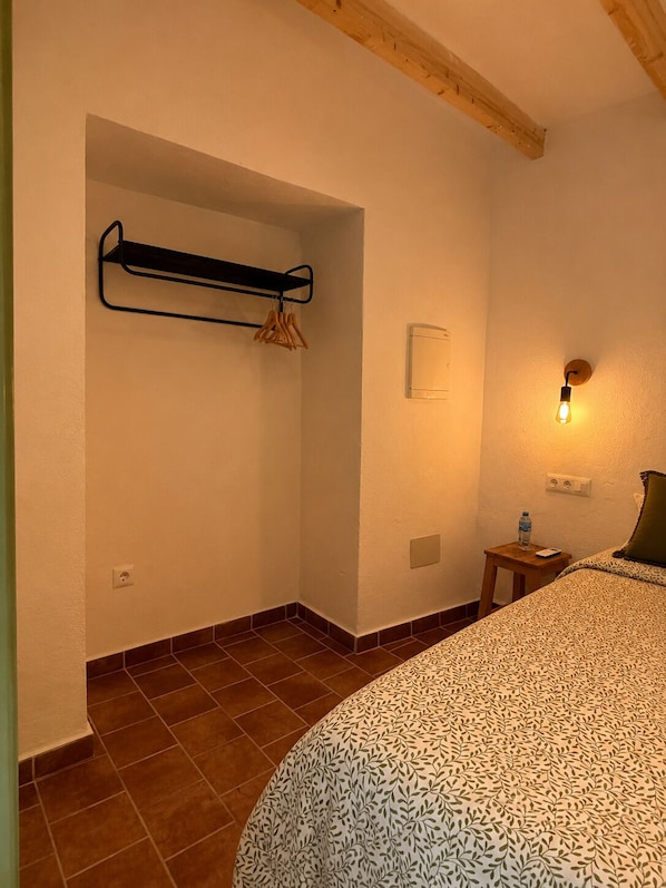 A bedroom with a bed, a hanging clothes rack, a wall-mounted light, a small table with a bottle, and a power outlet.