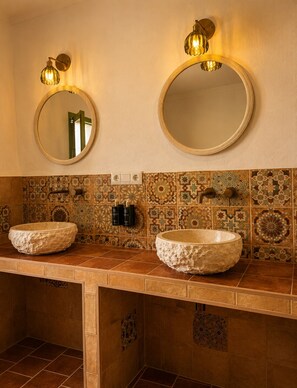 Une salle de bain avec deux miroirs ronds, un pare-chocs en carreaux de couleur beige et un meuble-lavabo en bois doté de deux grands vasques en pierre.