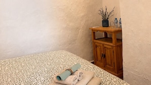 Une chambre à coucher avec une couvre-lit à motifs, une serviette enroulée et une petite armoire en bois ornée d’une plante.