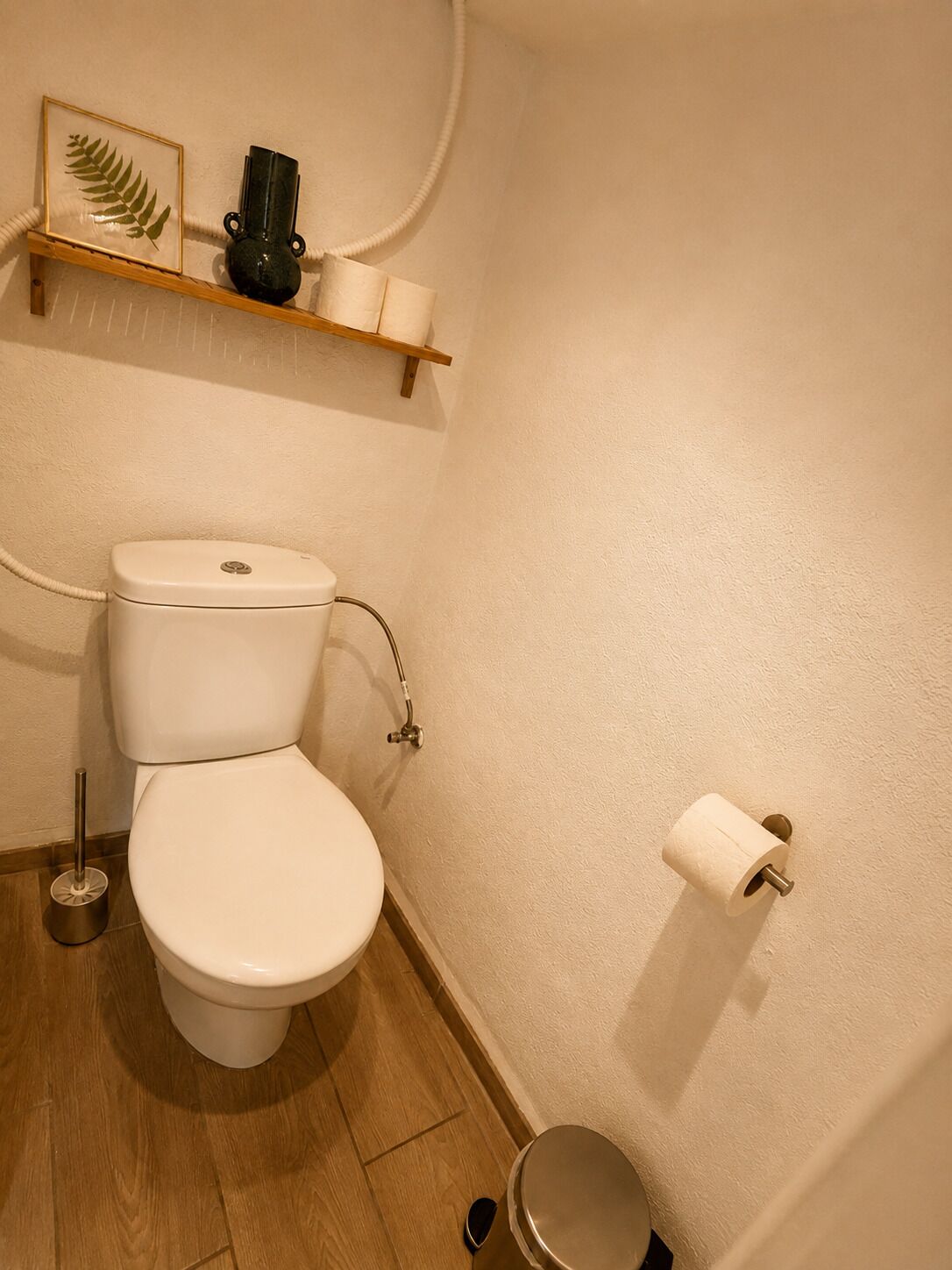 Une salle de bain avec des toilettes, une étagère ornée d’un cadre contenant une feuille, un vase et un porte-rouleau de papier toilette.
