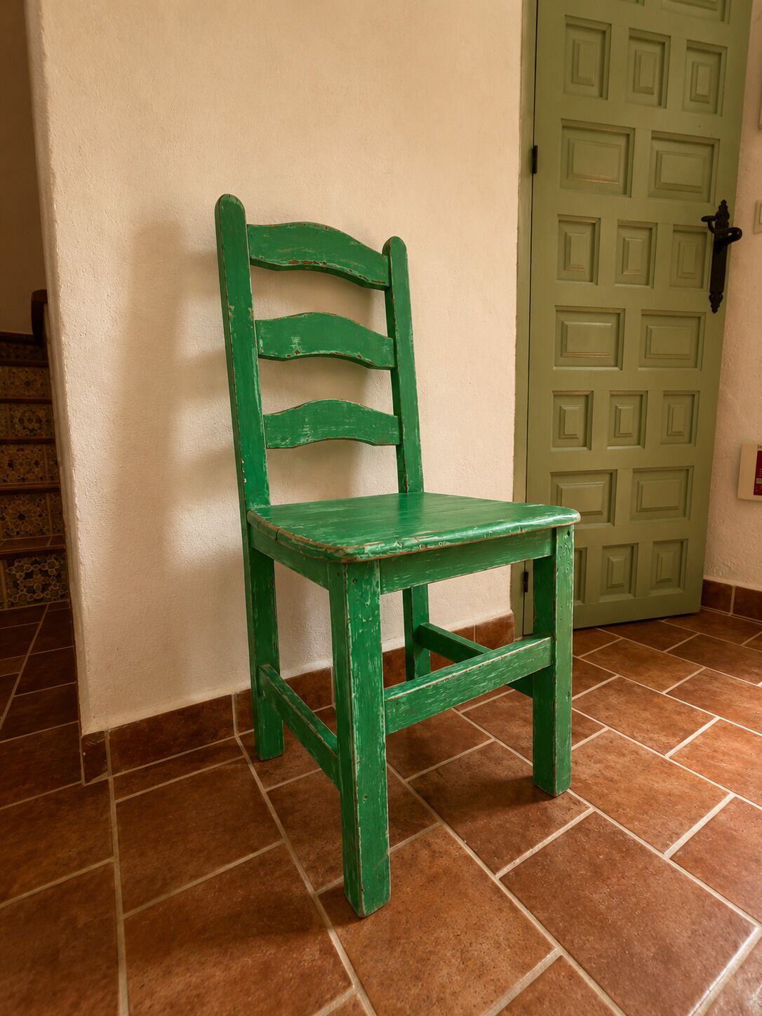 Een groene houten stoel met een eenvoudig ontwerp, geplaatst voor een muur met een groene deur op de achtergrond.