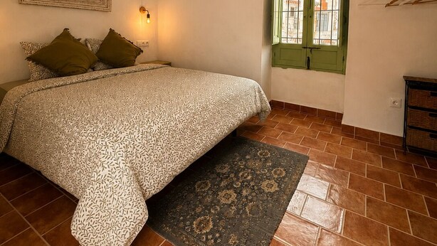 Ein Schlafzimmer mit einem gemusterten Bettüberwurf, einer hängenden Deckenleuchte, einem grünen Fenster und einem gefliesten Boden.