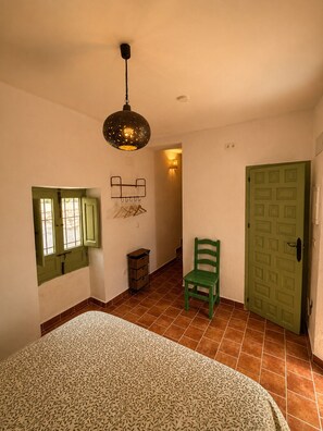 Une pièce avec une couvre-lit à motifs, une chaise verte, une porte verte, un porte-manteau fixé au mur et un luminaire suspendu.