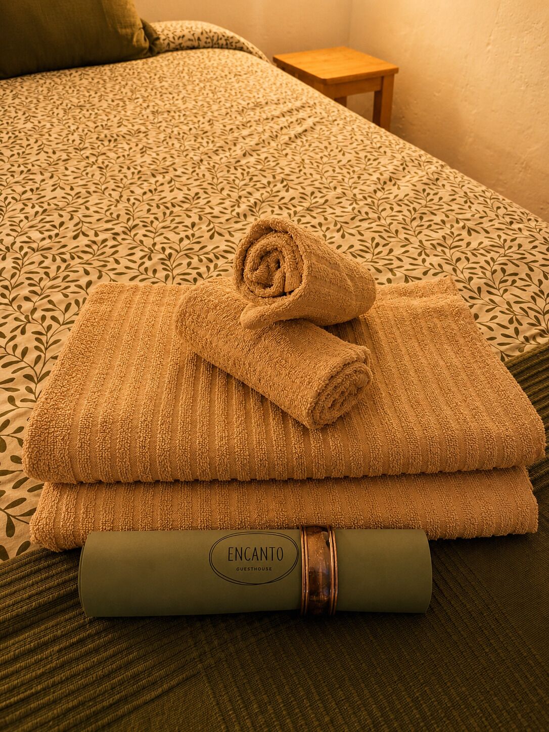 Un letto rifatto con una coperta a motivi, un asciugamano arrotolato e un oggetto avvolto con la scritta 'ENCANTO GUESTHOUSE'.