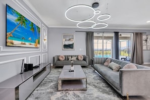 A modern living room with a flat-screen TV, a grey sofa, a coffee table, and a large window with a view. - Luxury 10BR Windsor Island Villa | 24 | Private Pool | Spa | Game Room |Theater (Davenport)