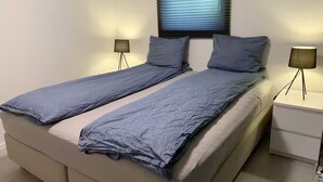 A double bed with a grey blanket, two bedside lamps, and a window with blinds.